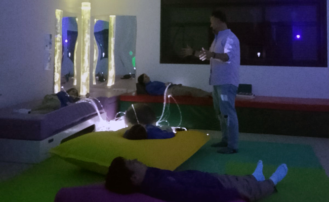 Tres chavales tumbados en distintos sitios de la habitación, con luces leds y escuchando al hombre que hay de pie en frente de ellos.