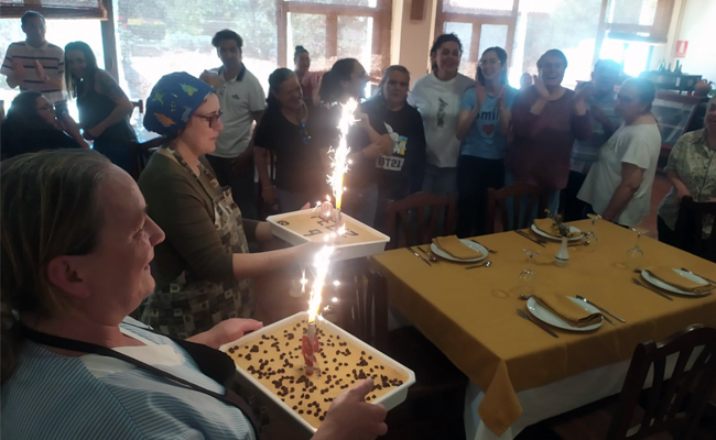 Dos mujeres llevan dos tartas de cumpleaños y están rodeadas de mucha gente para celebrar el noveno aniversario de la residencia de Cortegana.