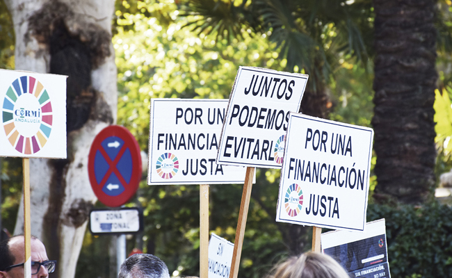 Fotografía de una protesta con varias pancartas en primer plano que exigen una financiación justa. Los carteles contienen mensajes como 'Juntos podemos evitarlo' y 'Por una financiación justa'. En el fondo, se observan árboles, una señal de tráfico de prohibido estacionar y un ambiente urbano.