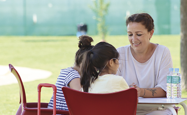 Mujer sonriente acompaña a dos niñas en una mesa al aire libre. La escena transmite un ambiente de apoyo e inclusión, representando la atención a menores del sistema de protección en Sevilla.