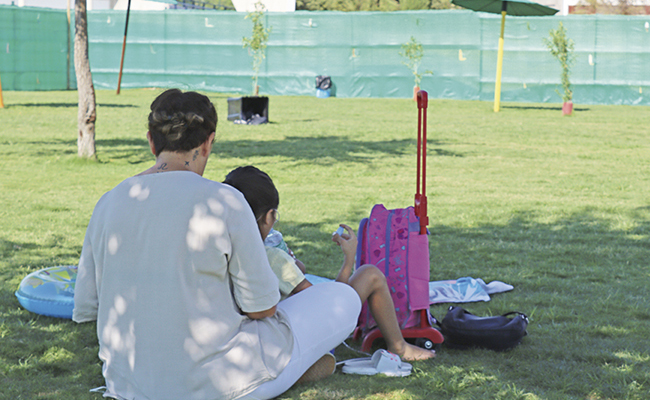 Mujer acompaña a una niña mientras están sentadas en el césped de un área verde. Junto a ellas, una maleta rosa y objetos personales sugieren un momento de cuidado y atención. La imagen representa el apoyo a menores del sistema de protección en Sevilla.